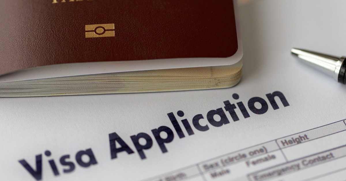 A close-up of a brown passport, a black pen, and a white piece of paper that says "Visa Application."