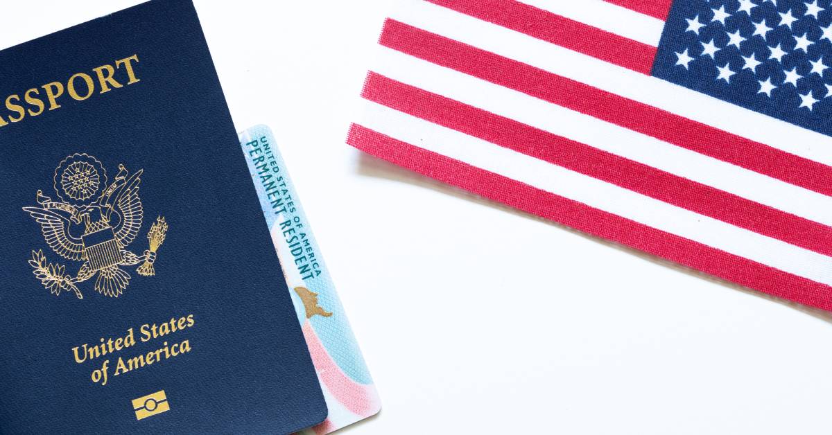 A close-up of a United States of America passport, a Permanent Resident Card, and an American flag on a white background.