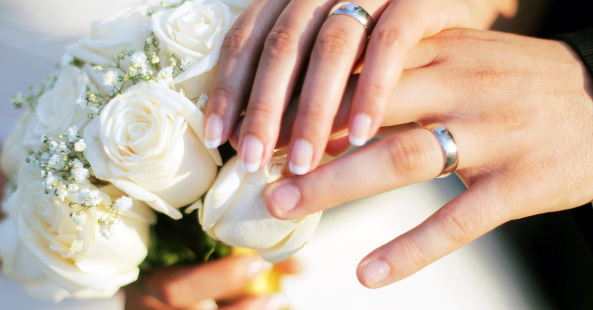 A bride and a groom wearing silver wedding rings on their left ring fingers. The bride holds a bouquet of flowers.