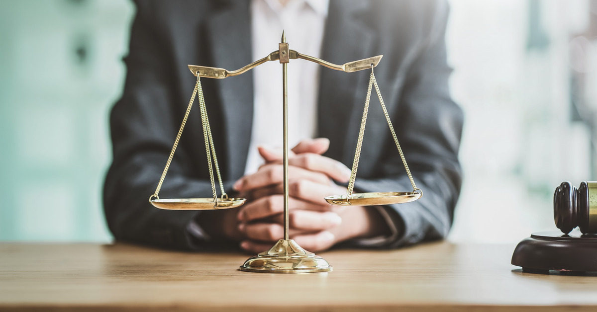 A man in a suit seated at a table with his hands folded. The scales of justice and a judge's gavel are on the table.