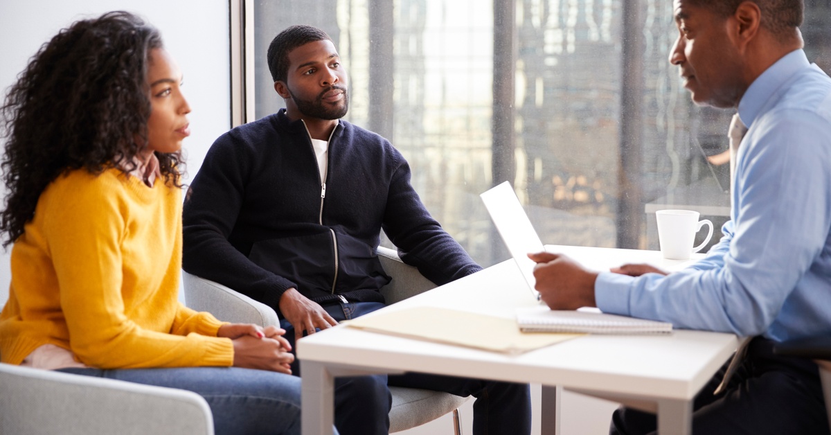 A man in professional attire sitting at a white desk and speaking to a man and a woman sitting across from him.