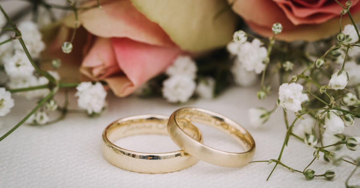 A close-up of two gold wedding rings stacked together. There are roses and baby's breath in the background.