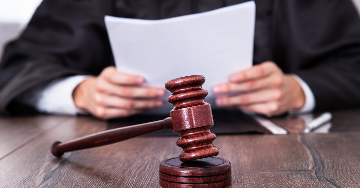 A judge is holding a thin stack of papers. A wooden gavel is sitting on the table in front of them.