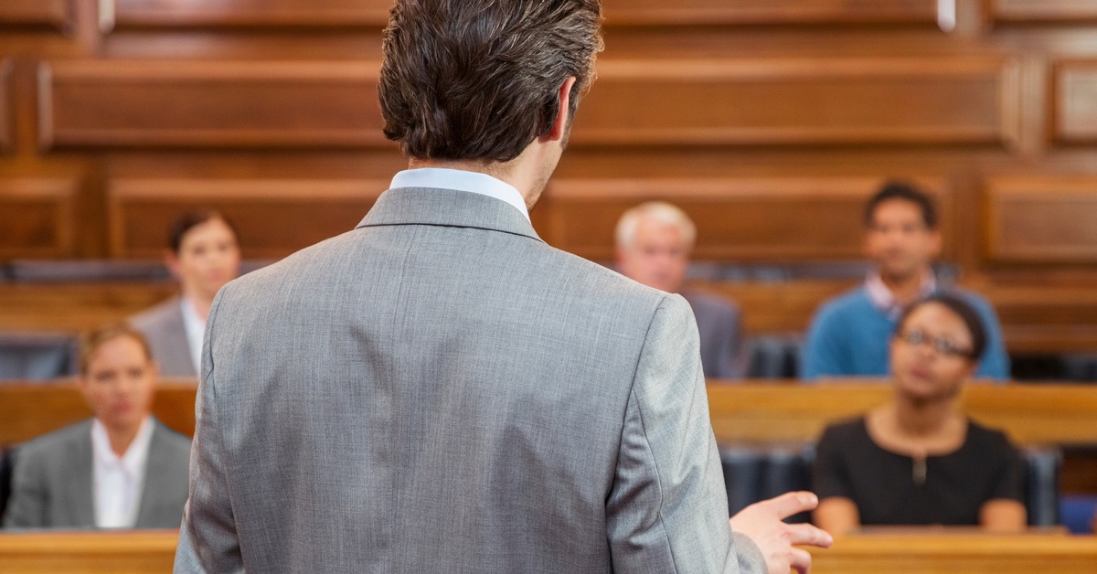 A male lawyer in a gray suit is addressing a jury intently listening to him inside a courtroom.