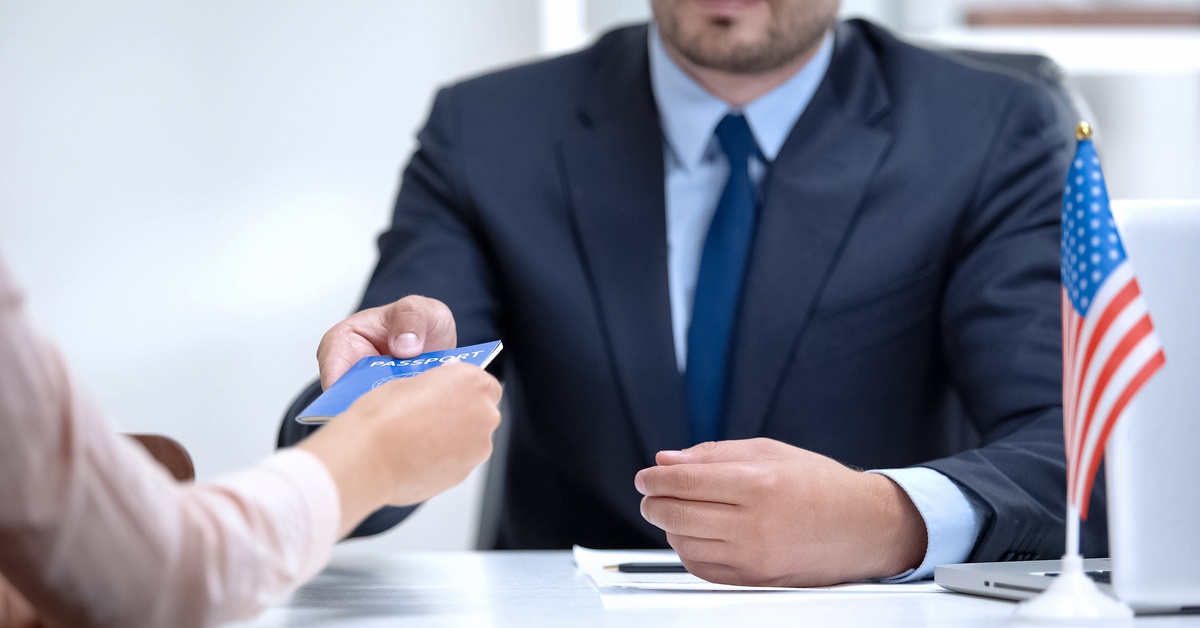 A man in a formal blue suit hands a passport to another person across a table with an American flag and laptop on it.