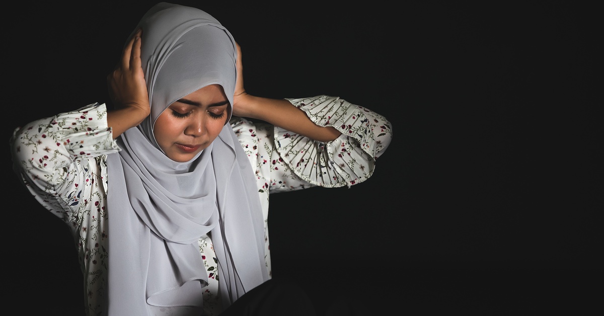 A young woman wearing a gray hijab and white floral shirt is covering hear ears while looking distressed.