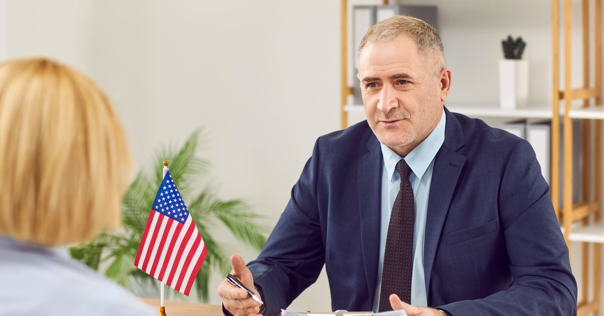 A man in a navy suit holding a pen and clipboard while talking to another person. There is an American flag on his desk.