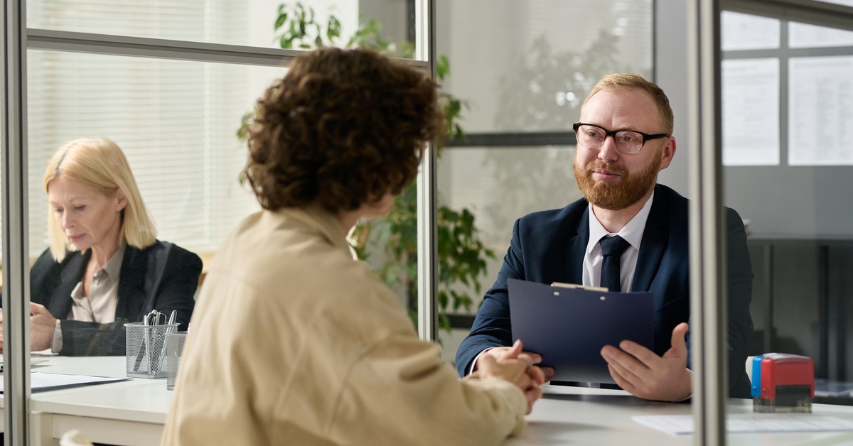 A man sitting in a cubicle, smiling warmly while holding a clipboard. He is chatting with someone sitting across from him.