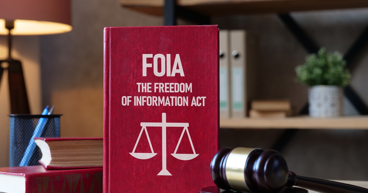 A red book with "FOIA" and the scales of justice on the cover stands on a table next to a gavel and eyeglasses.