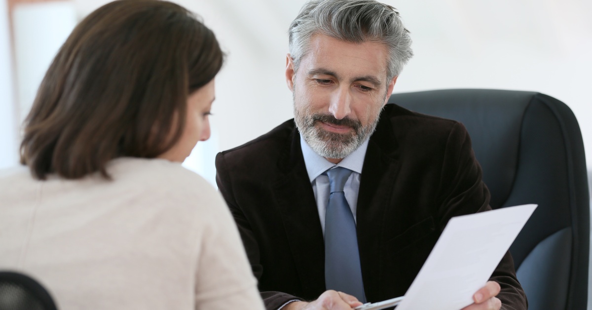 An older man wearing a black suit points to a document with his pen while smiling at a woman sitting across from him.