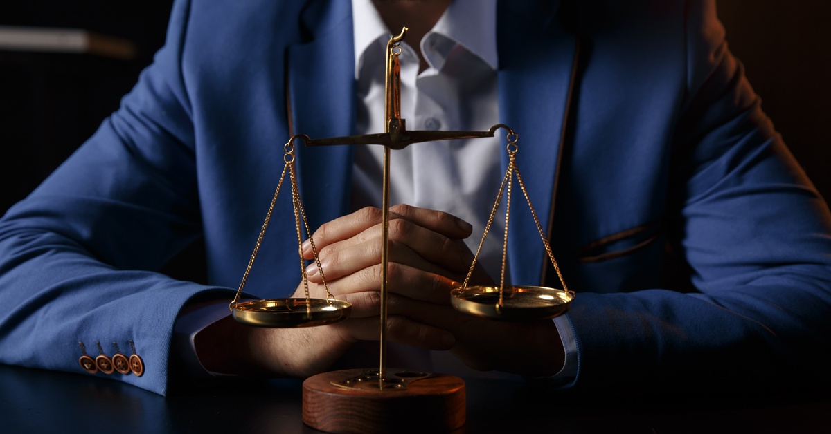 A person wearing a blue suit sits with their hands crossed behind a gold-plated and wooden scales of justice.