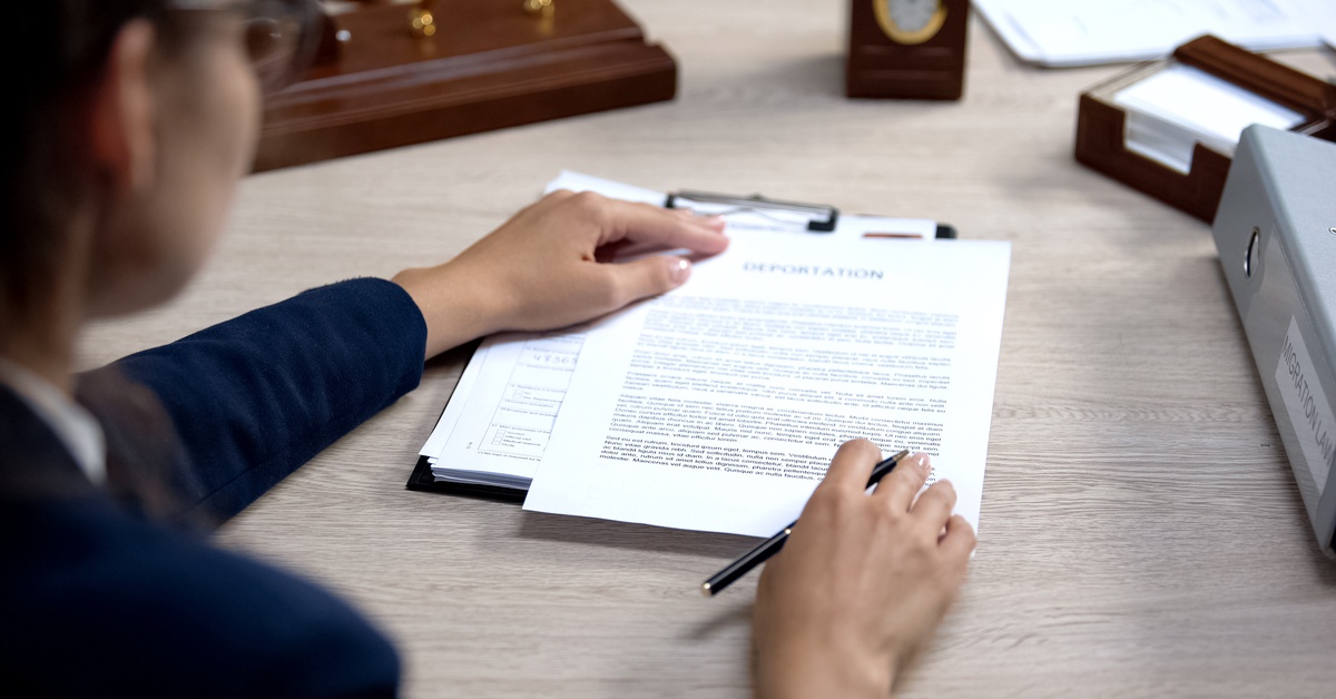 A person wearing glasses looks over documents on a clipboard that says "deportation" while holding a black pen.