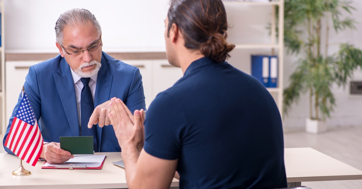 A person pleads with his hands while an immigration officer sits across him reviewing his documents.