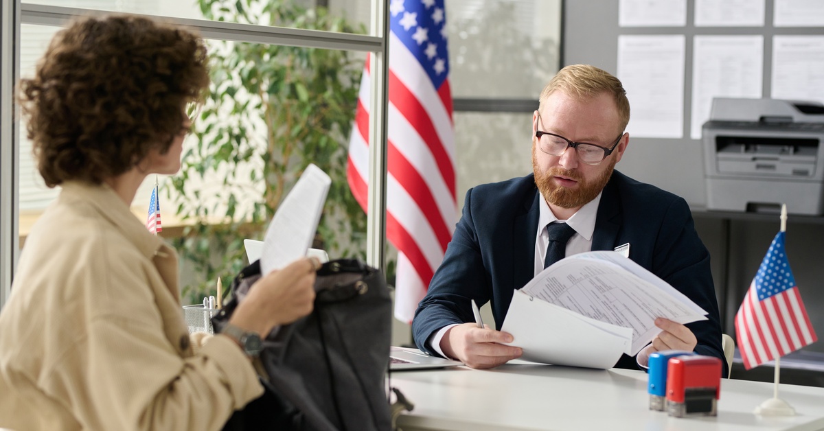 A male U.S. Immigration officer is flipping through documents while another person sitting across them holds a bag.