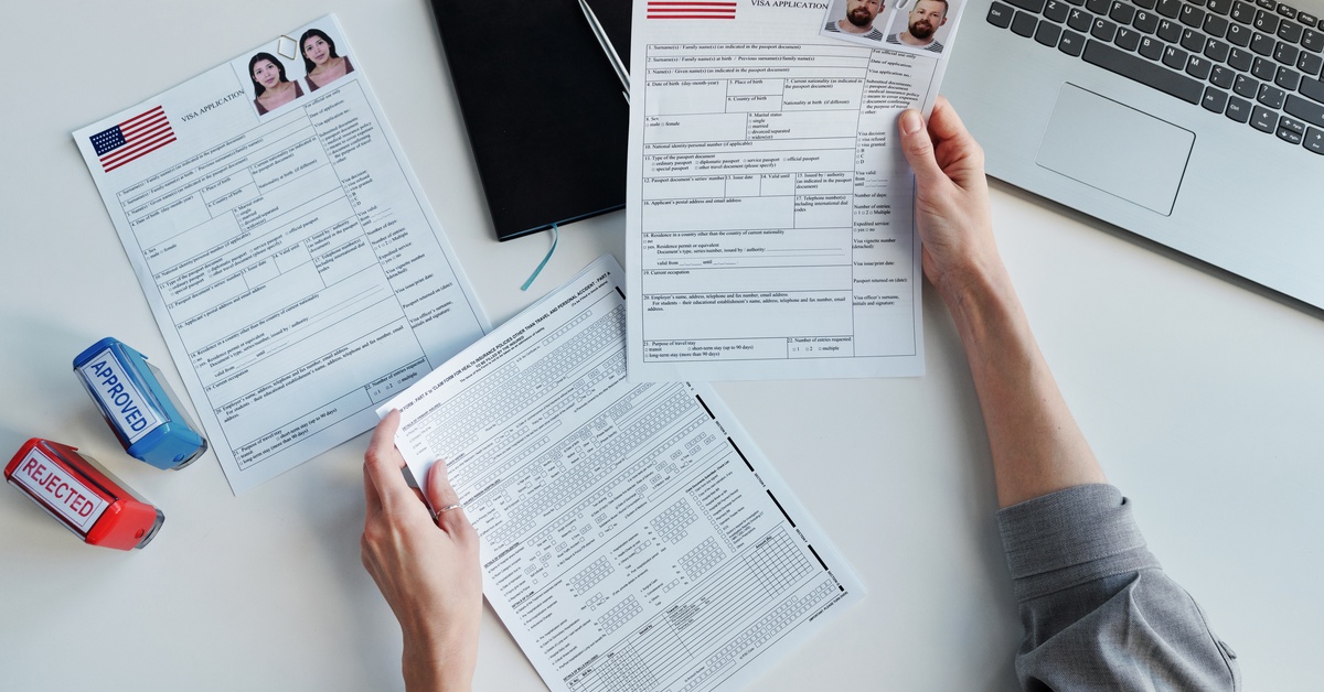 Top-down shot of someone holding visa applications, next to a laptop, an approved and rejected stamp, and a notebook.