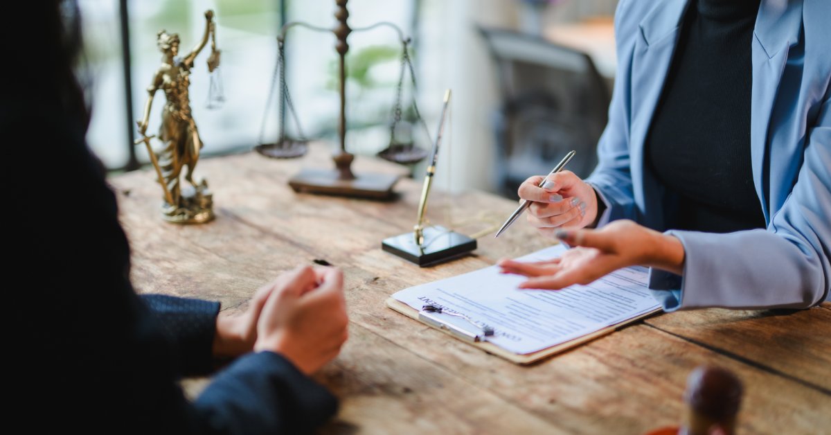 A person talks to another person sitting across them. On the table is a clipboard, a gold pen, and the scales of justice.