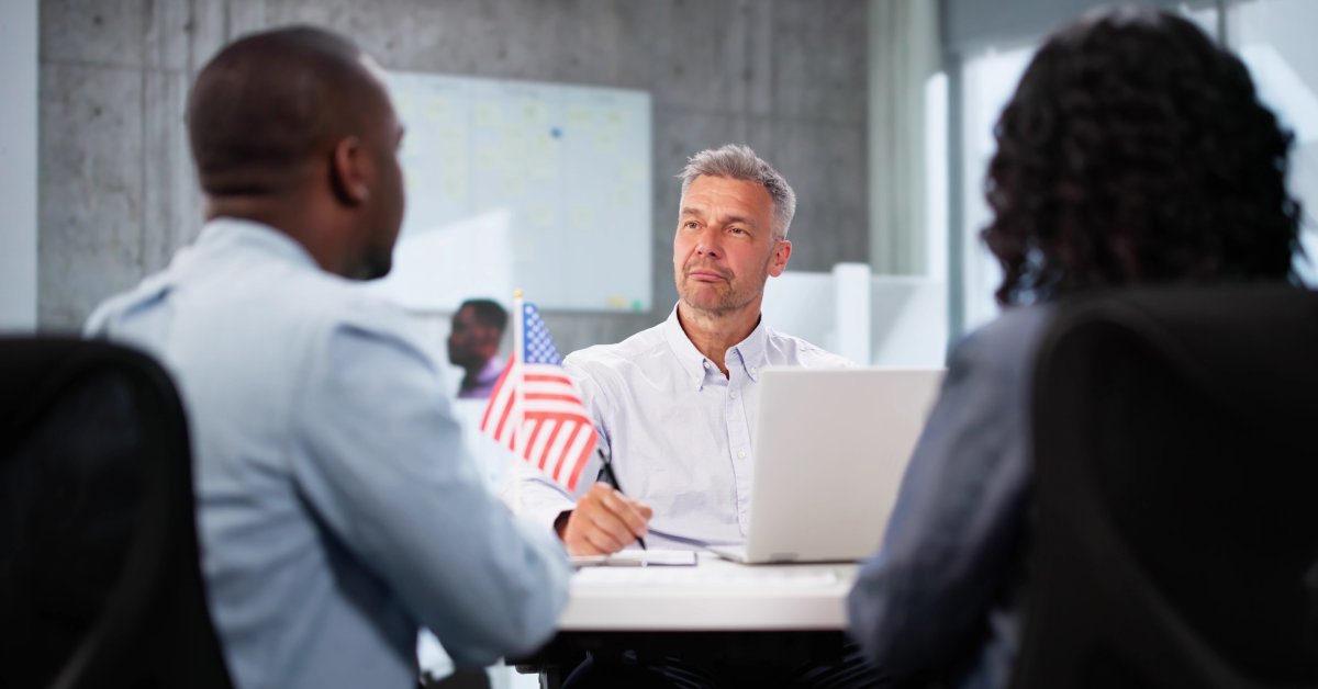 A man wearing a white shirt sits across two other people. On the table is the American flag and a white laptop.