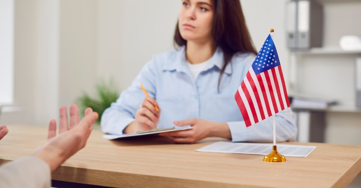 A woman listens to a person sitting across her while jotting down notes on a clipboard next to the American flag.