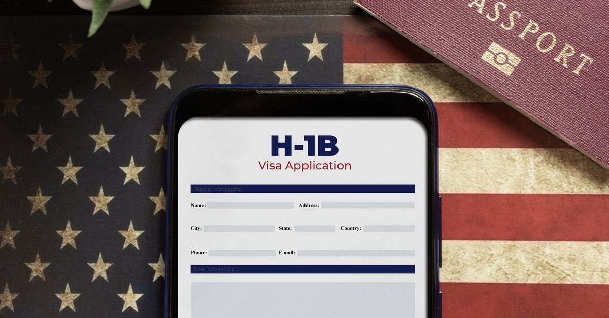 A phone with the H-1 Visa Application pulled up is sitting on top of the USA Flag next to a red passport.