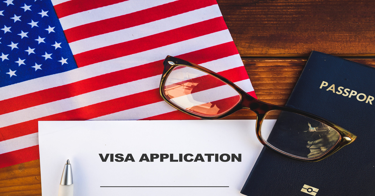 The flag of the United States, a visa application document, a passport, and eyeglasses sit on top of a table.