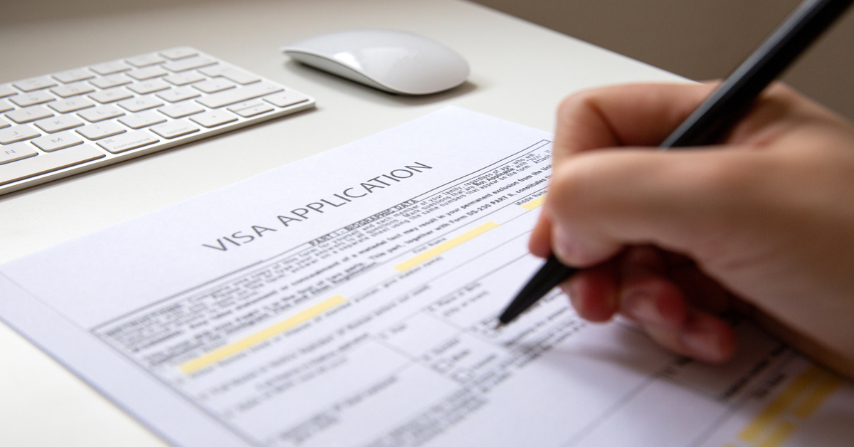 A person writes on a visa application document sitting on top of a white table next to a keyboard and mouse.