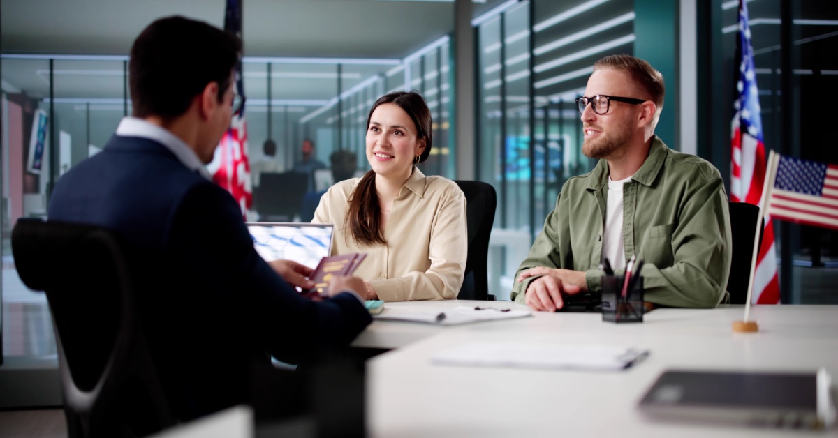 A man wearing a navy blue suit holds two passports while sitting across a man and a woman, surrounded by U.S. Flags.