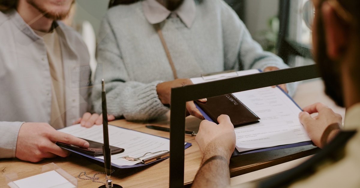 Two people stand next to an administrative cubicle, smiling, as they hand over both their passports and documents.