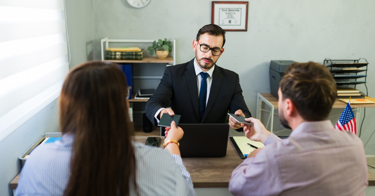 A lawyer wearing a black suit, blue tie, and eyeglasses, takes the passports of a couple sitting across the table.
