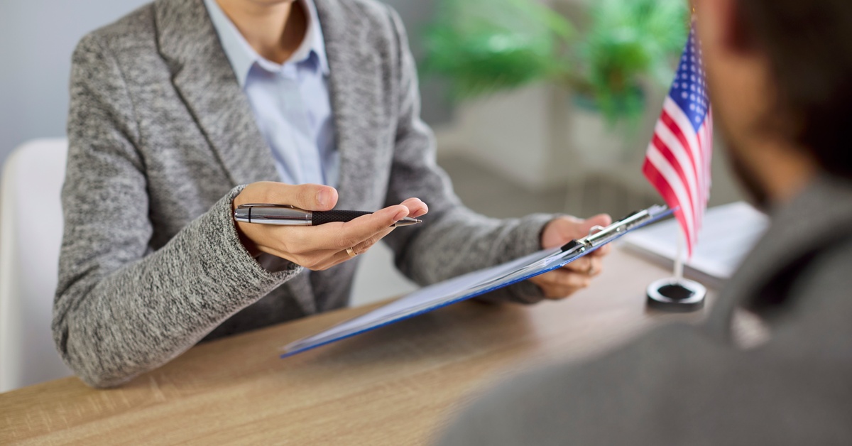 A lawyer wearing a gray suit holds a pen with one hand and a clipboard with the other while sitting across a client.