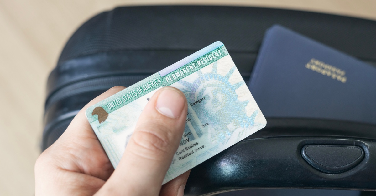 A person holds a permanent resident card, covering the information. Behind it is a black travel suitcase.