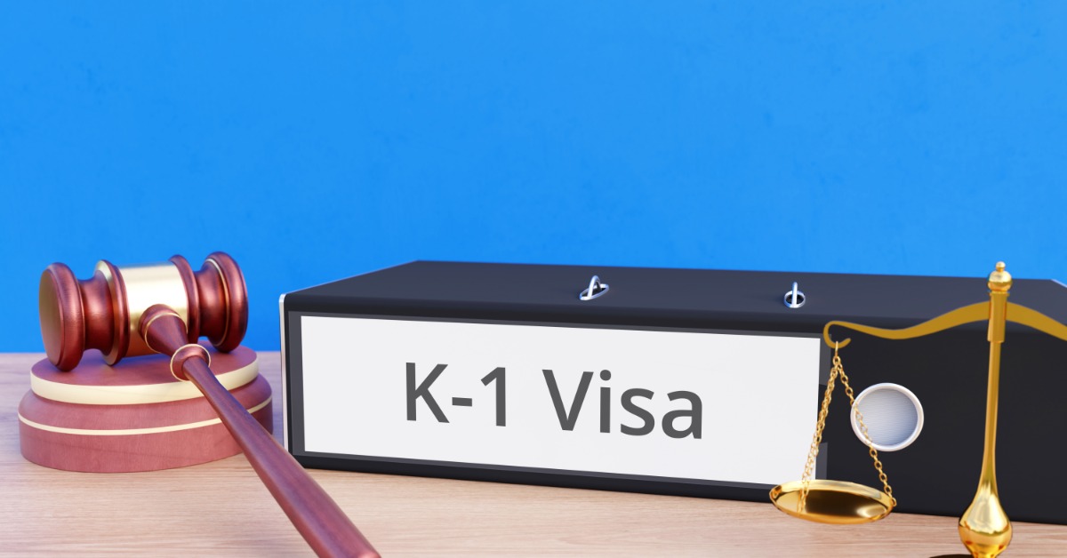 A thick black binder labeled "K-1 Visa" sits on top of a table next to a brown gavel and a golden scales of justice.