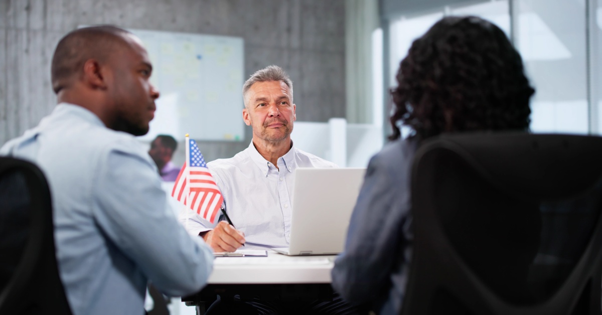 Two people sit across a man wearing a white shirt for a visa interview. On the table is the U.S. flag and a laptop.