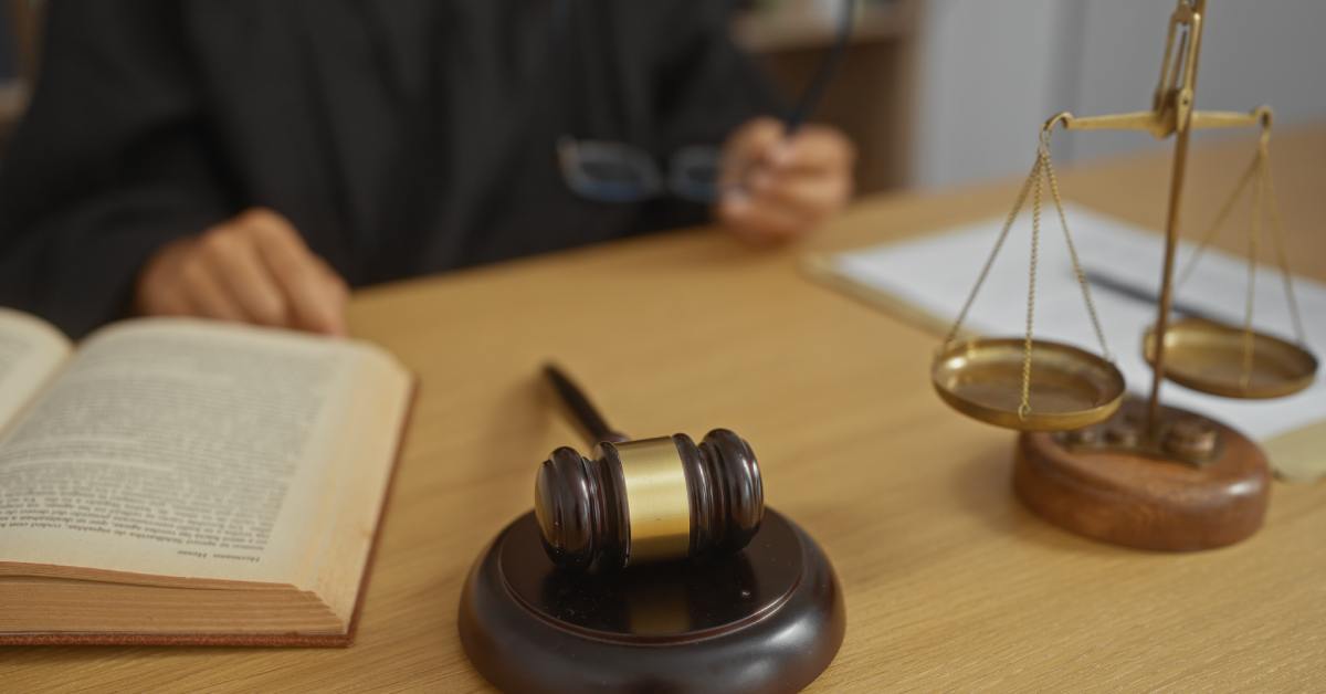 Judge sits next to a legal book, gavel and scales on a desk, showing courtroom decisions during an indoor hearing.