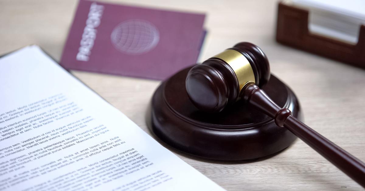 A red passport sits beside a brown gavel, sound block, and immigration documentation on top of a wooden table.