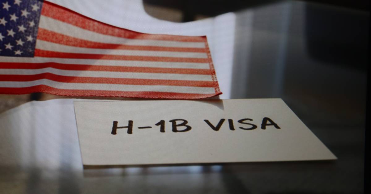 An illustration of an H-1B visa document next to a U.S. flag on top of a black glass table, reflecting shadows.