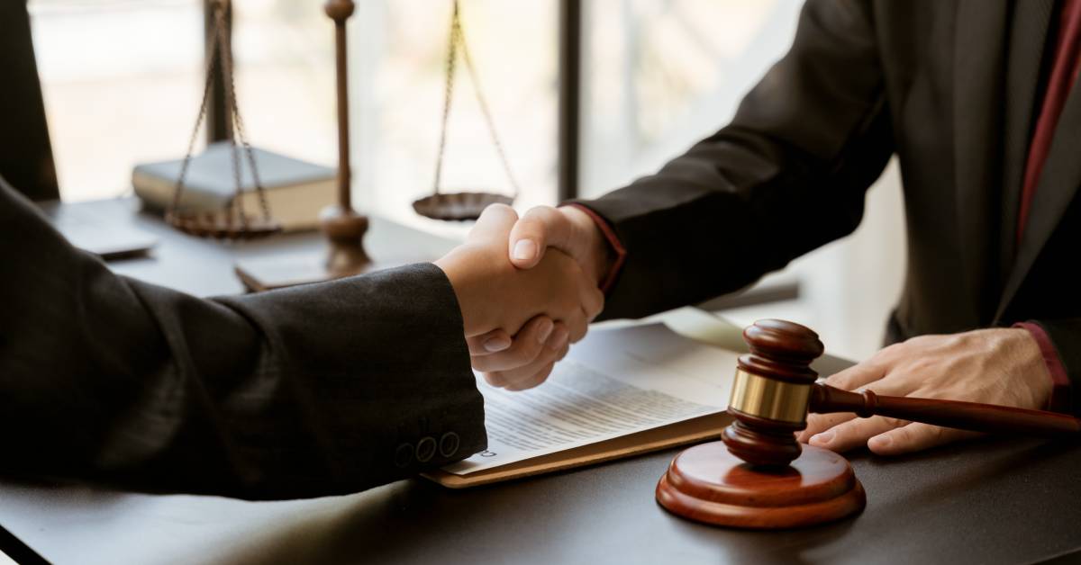 Two people shaking hands across a desk with a gavel, legal papers, and scales visible in the background.