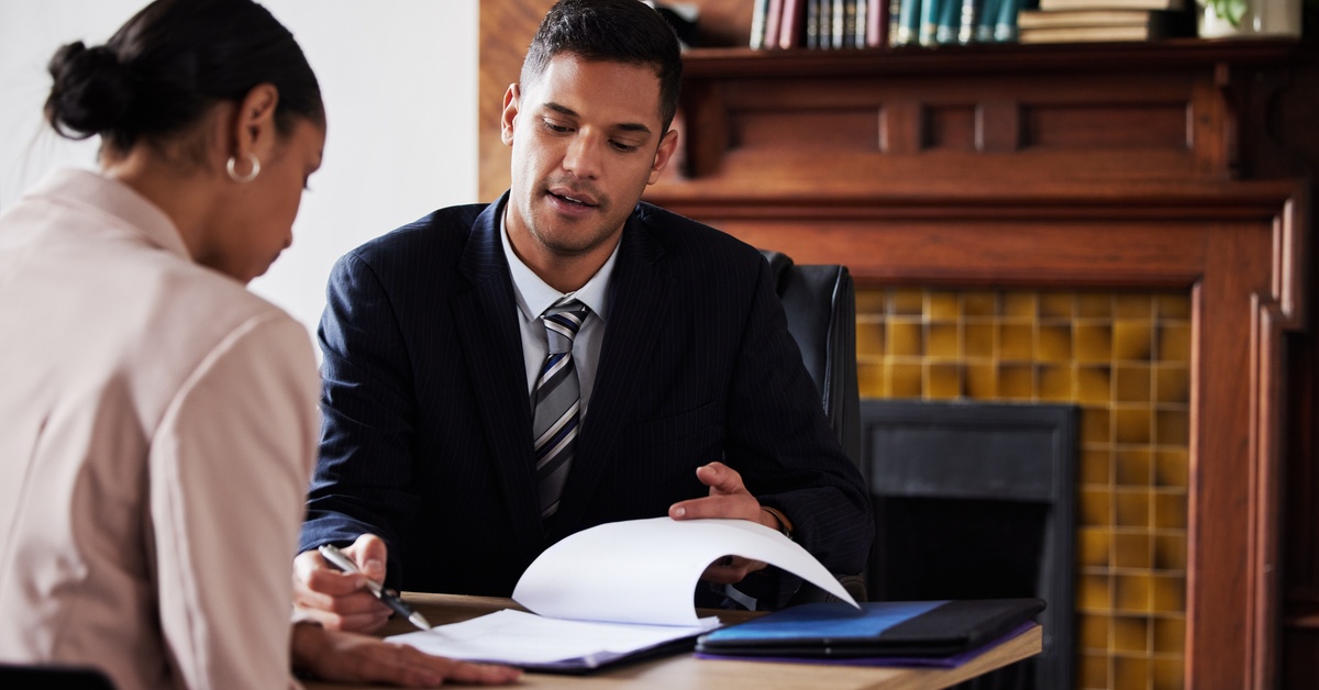 A man in a suit and tie uses a pen to point at documents. A woman sits across from him and listens attentively.