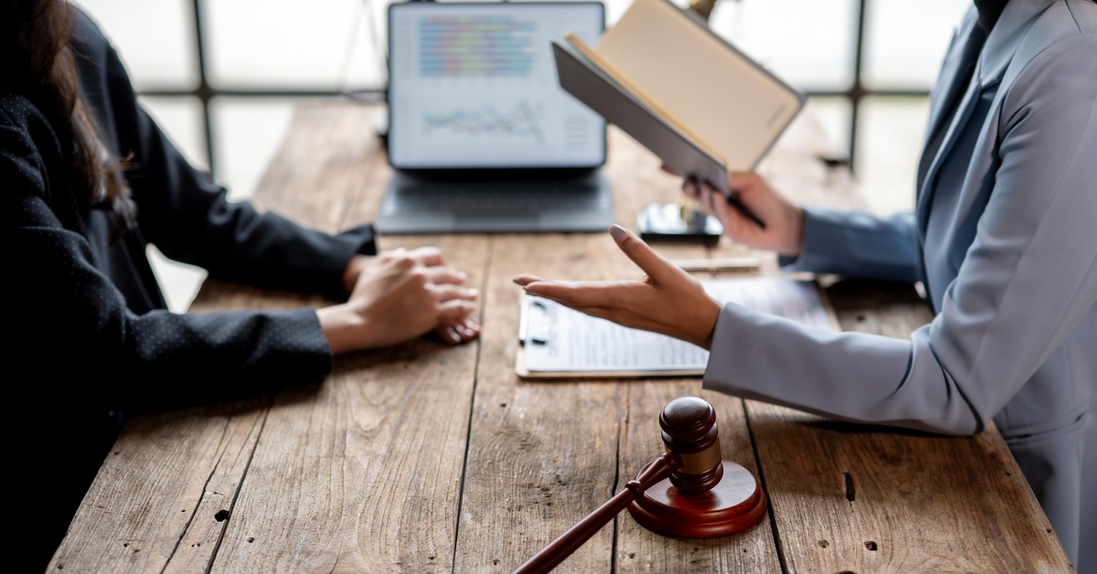 A woman wearing a light blue blazer holds a book and speaks to another woman. They are sitting at a wooden table with a gavel.