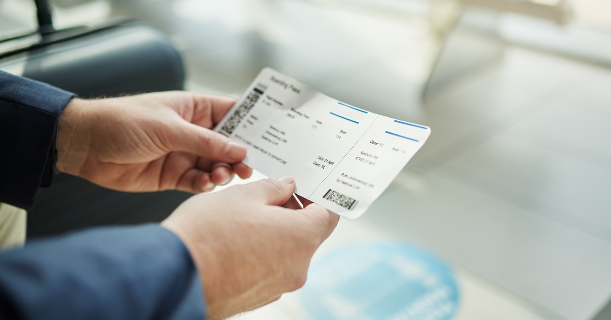 A person in a blue long-sleeve shirt sits holding a plane ticket. A blue suitcase is to their left.