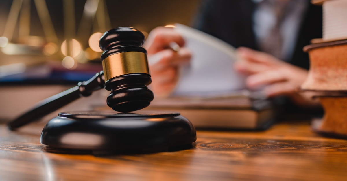 A gavel is in focus as a person reviews papers, surrounded by books in the background.