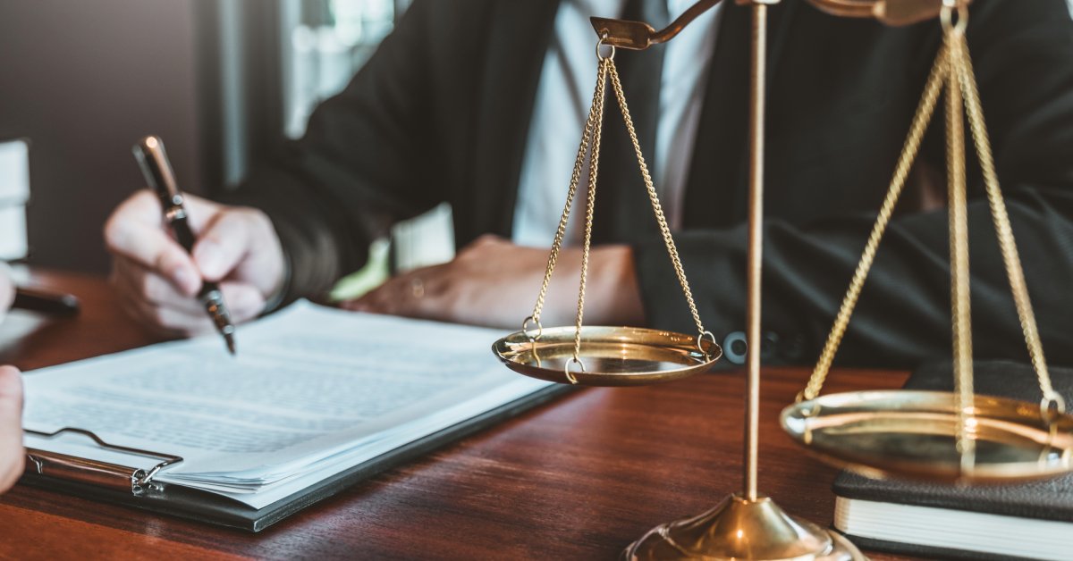 A person in a black suit writes with a pen on documents on a clipboard next to a gold scales of justice.