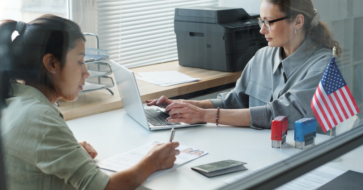 Two women sits across each other as one writes on a paper and the other types on a laptop sitting on a table.