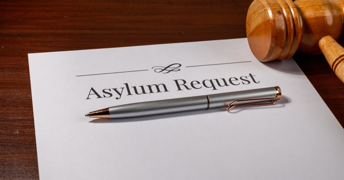A document that says "Asylum Request" sits on top of a wooden table next to a gavel and a silver ballpoint pen.