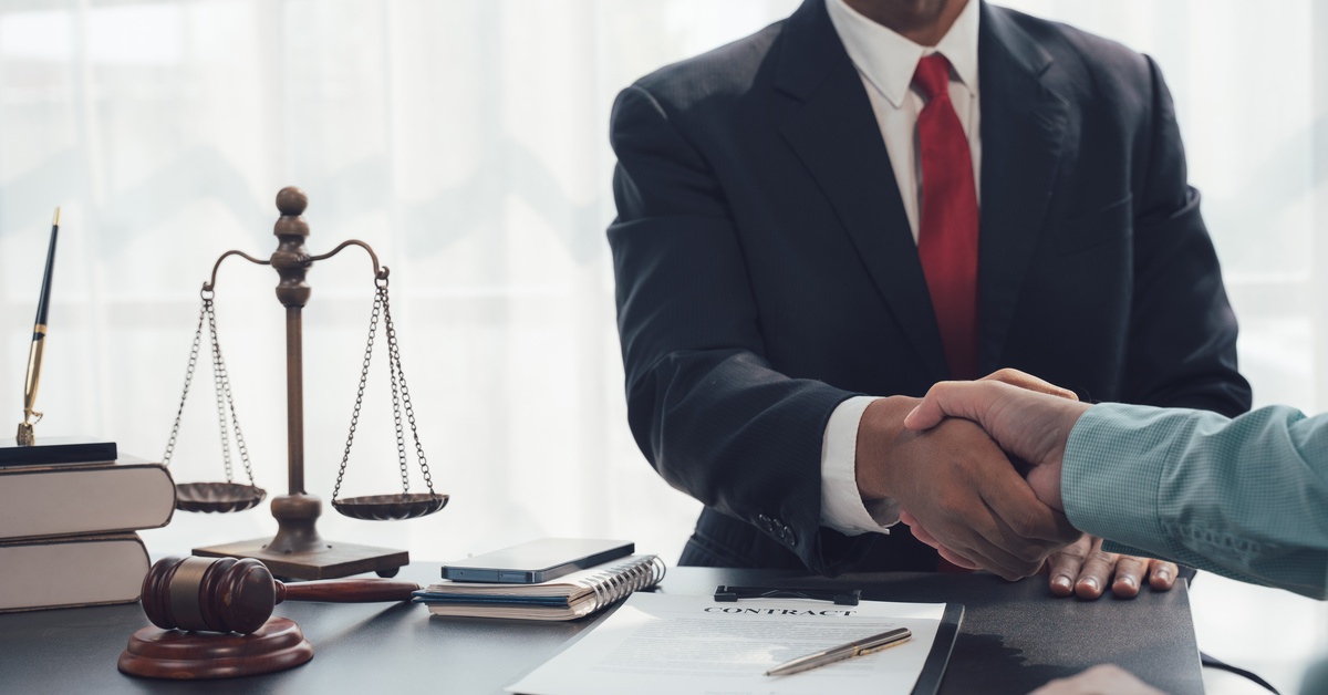 Two people shake hands across each other. In between is a table with the scales of justice, documents, and gavel on top.