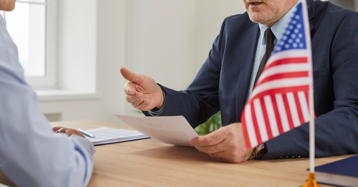 A man wearing a navy suit and glasses holds a document and sits across a woman. A small American flag stands next to them.