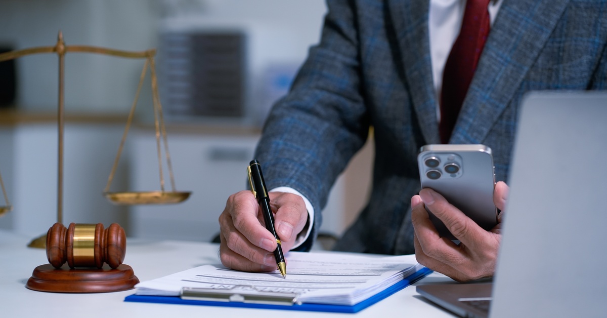 A person in a navy suit signs documents with a pen and holds a smartphone. On the table is a scale of justice, gavel, and laptop.