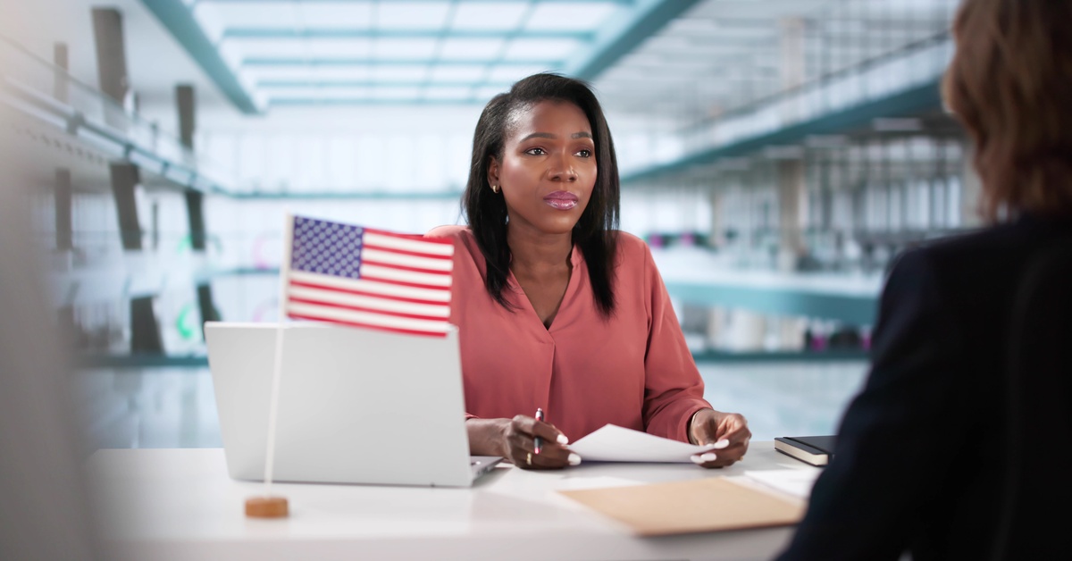A woman wearing a coral blouse sits across another person. Beside her is a laptop and the American Flag.