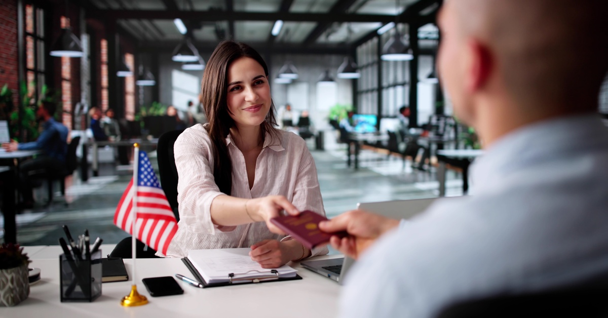 A woman wearing a pink shirt handing over a passport across the table to another person. On the table is a US Flag.