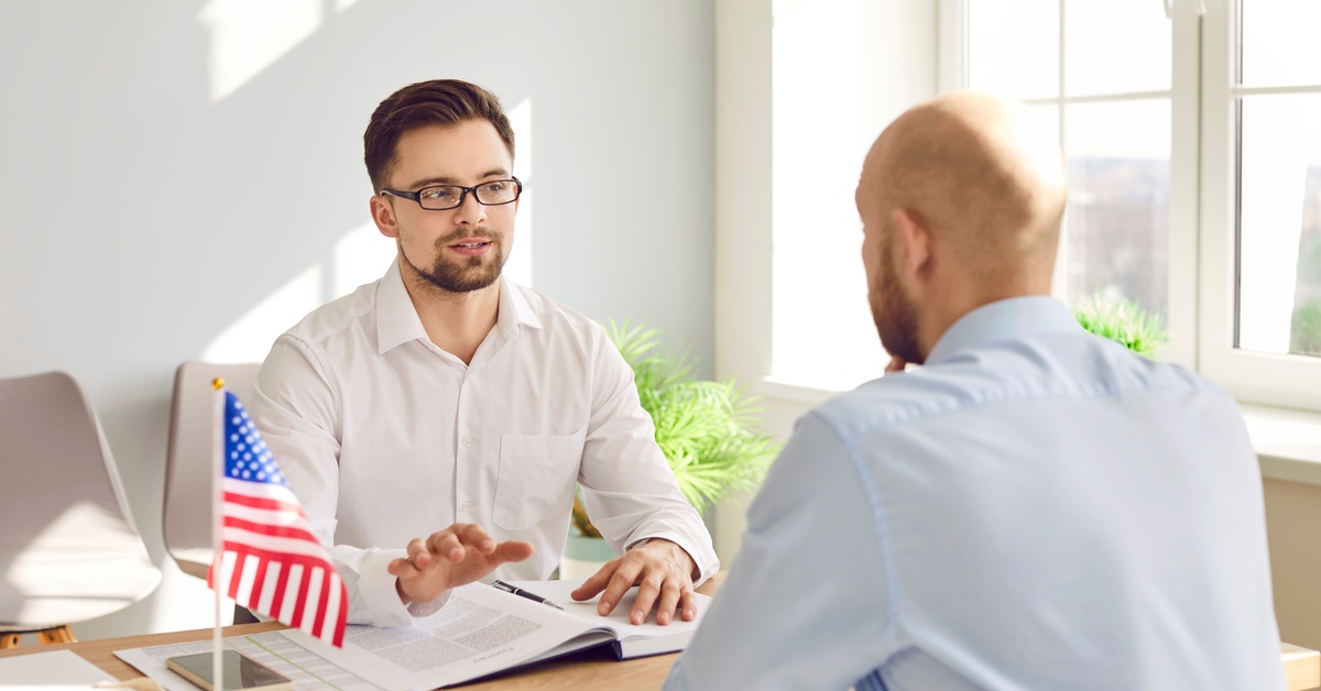 A man wearing glasses is talking to a person sitting across him. On the table is the American Flag and documents.