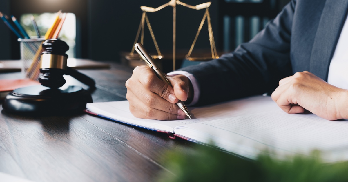 A person is writing on a notebook with a gold pen. Next to them is a gavel and behind is the scales of justice.